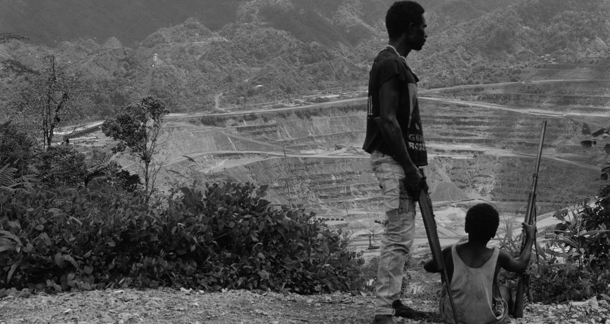 Black and white photograph of a Bougainvillian man holding a gun looking into the dormant Panguna mine. A child sits to his right with a gun.